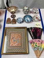 Full lot overview showing all oriental items including porcelain dishes, Tibetan figurines, framed artwork, and fans on a white tablecloth.