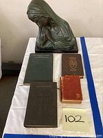 Religious lot with praying Madonna bronze bust sculpture and four religious books on white fabric surface, labeled with number 102.