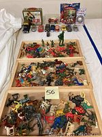 Overall view of the lot showing wooden trays with assorted plastic toy soldiers, cowboys, animals, and other figures spread out as well as several die-cast cars and the G.I. Joe tin behind.
