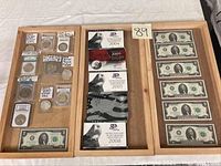 Three wooden trays showing slabbed coins in left tray, boxed state quarter sets in middle, and $2 bills in right tray.
