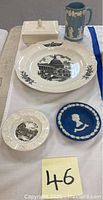 Full view of all items in the lot laid on white cloth: large and small Capitol building plates, blue Jasperware medallion, covered dish with lid, small pitcher.