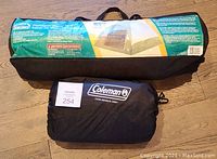 Two bags on wooden floor. One green and black bag containing the Pentagon Dome 4 person tent, and one black bag labeled Coleman containing a twin air mattress.