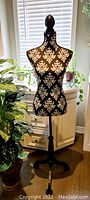 Full view of black and white damask patterned dress form mannequin standing near window with plants.