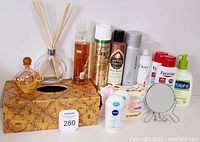 Photo showing an assortment of bathroom and personal care items including perfume bottle, reed diffuser, body spray, hair sprays, deodorants, lotion bottles, and a small standing mirror. Includes decorative bee-themed tissue box as a base.