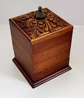 Closed view of wooden box showing carved floral design on top with brass finial handle.