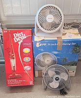 Dirt Devil cordless stick vacuum in original box, white Honeywell desk fan on top, Lakewood gray metal blade fan in front, Super oscillating fan box behind.