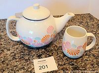 Photo of white ceramic teapot with colorful fan-shaped pattern and matching cup on granite countertop with a lot number card.