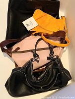 Four bags arranged in a pile showing size and color variety: black tote, yellow small bag, beige and brown handbag, black handbag with white stitching.