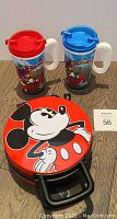 Mickey Mouse waffle maker with red lid and two Disney plastic cups with lids and handles (red and blue).