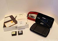 Photo showing Nintendo DS XL original box, 2 game cartridges labeled "My Spanish Coach" and an unidentified second game, docking station, and red-black carrying case.