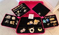 Pink case open showing various compartments filled with costume jewellery including brooches, earrings, and rings.