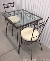 View of vintage metal table with glass top and two matching chairs with cream cushions showing condition and style.