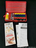 Open red plastic case containing five colorful sharpening stones, honing oil bottle, mounting clamp, and adjustment rods.