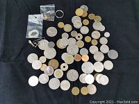 Wide view of assorted vintage and foreign coins scattered on black background, showing variety of sizes, colors, and some coins with central holes. Includes small plastic bags with keys and rings.