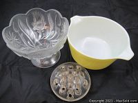 Three items on a black background: vintage yellow Pyrex bowl with white interior, pressed glass floral bowl on pedestal, and round glass flower frog with holes.