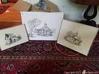Three black and white framed art prints placed on a carpet showing detailed drawings of rustic buildings
