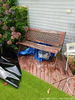 Front and partial side view of the bench placed outdoors on wooden deck with plants nearby. Shows overall structure, steel lattice back and sides, and heavily weathered wooden seat slats.