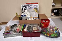 Box and containers holding assorted arts and crafts materials including paints, brushes, an art book, craft foam, and glue.