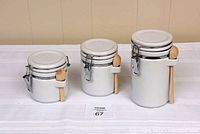 Photo showing three white ceramic canisters of varying sizes with locking metal lids and attached wooden spoons on a white surface.