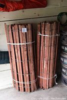 Two rolled bundles of cedar snow fencing leaning against a wall inside a garage area. Each bundle is about 25 feet in length and secured by white plastic ties.