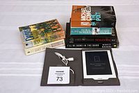 Photo showing Kobo eReader in brown protective case with white charging cable, six books stacked to the side on white bedspread.