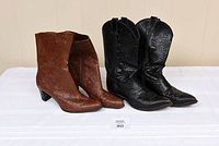 Photo shows two pairs of ladies cowboy boots side by side on a white surface against a cream wall. The left pair is brown with embossed design and low angled heels. The right pair is black leather with Western stitching and mid-height heels, visible heel wear.