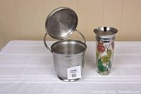 Aluminum ice bucket open next to martini shaker with cocktail graphics on table.