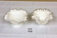 Two white milk glass bowls with ruffled edges and silver trim, one on a pedestal and one without, displayed on white fabric.