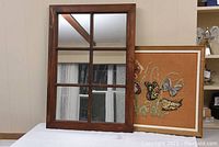 Window pane mirror framed in stained wood leaning against a brown frame with crewel work on a white surface