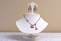 Photo showing rose gold color necklace with round triple AAA Zirconium centerpiece and matching dangle earrings on a white display stand against a neutral background.