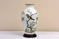 Full front view of the white porcelain vase featuring one bird on flowering branch with green leaves and pink flowers. 24 karat gold rim visible, vase resting on a wooden base.