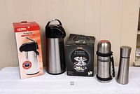 Photo of a collection of four items: boxed Thermos vacuum pump coffee pot, Keurig carousel coffee pod holder in box, Starbucks thermos, and larger Arcosteel thermos, all stainless steel with black accents, standing against a beige wall on a table.