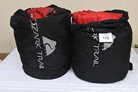 Photo showing two black cylindrical storage bags labeled 'Ozark Trail', each containing a red sleeping bag inside.