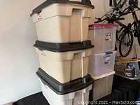 Stacked three large beige Rubbermaid Roughneck storage boxes with black hinged lids and three medium Sterilite clear containers with violet magenta lids in a garage setting.