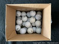 Top-down view of 25 assorted used golf balls in a cardboard box.