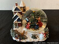 Photo of Christmas water musical globe showing snow-covered village house, snowman, deer figurine, and globe dome with children and adult decorating Christmas tree.