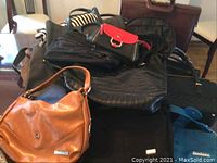 Photo showing a pile of bags including a large brown handbag, small black and red bag, and multiple black bags stacked on a chair.