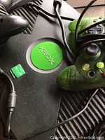 Close-up of original black Xbox console with green translucent wired Xbox controller resting on top with visible Xbox logo and cables.