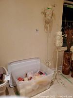 Plastic bin with clear lid containing assorted candles of various colors and sizes, positioned beside two tall white metal candle holders with scrollwork.