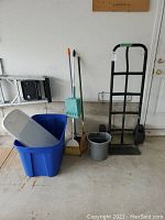 Full lot with dolly, blue storage bin with white lid inside, gray pail, broom, broom with orange handle tip, and green dustpan with handle standing on floor in garage.
