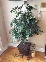 Full view of faux leafy plant in basket planter with metal stand and two ivory pillar candles on hardwood floor against beige wall.