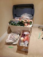 Large plastic bin with hinged lid containing assorted faux flowers with green leaves and peach-colored blossoms, and other assorted decorations. Next to bin is a cardboard box of assorted pillar candles and a clear bag of tissue paper.