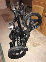 Folded black golf caddy with three rugged wheels placed on a basement floor near wooden cabinets.