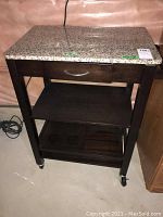 Full front view of a dark wooden kitchen cart with granite top and caster wheels on a basement floor.