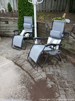 Two grey mesh fabric foldable gravity chairs with black metal frames placed outdoors on brick patio. Each has pillows and side drink trays.