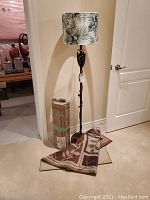 Floor lamp with green and white leafy tube lampshade next to a rolled carpet and a partially displayed carpet runner on the floor.