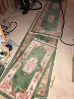 Photo of three matching wool carpet runners laid out on basement floor, showing green backgrounds with floral border patterns and fringed edges.