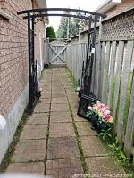 Front view of black metal garden arch with integrated lower gate, hanging planters and urn stands along a brick pathway