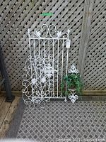 Full view of metal gate, plant holder with greenery, and two wall sconces against lattice background.