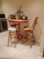 Full view of the round oak pub height table with 2 oak tall chairs and 1 fabric seat stool grouped around it in corner of room.